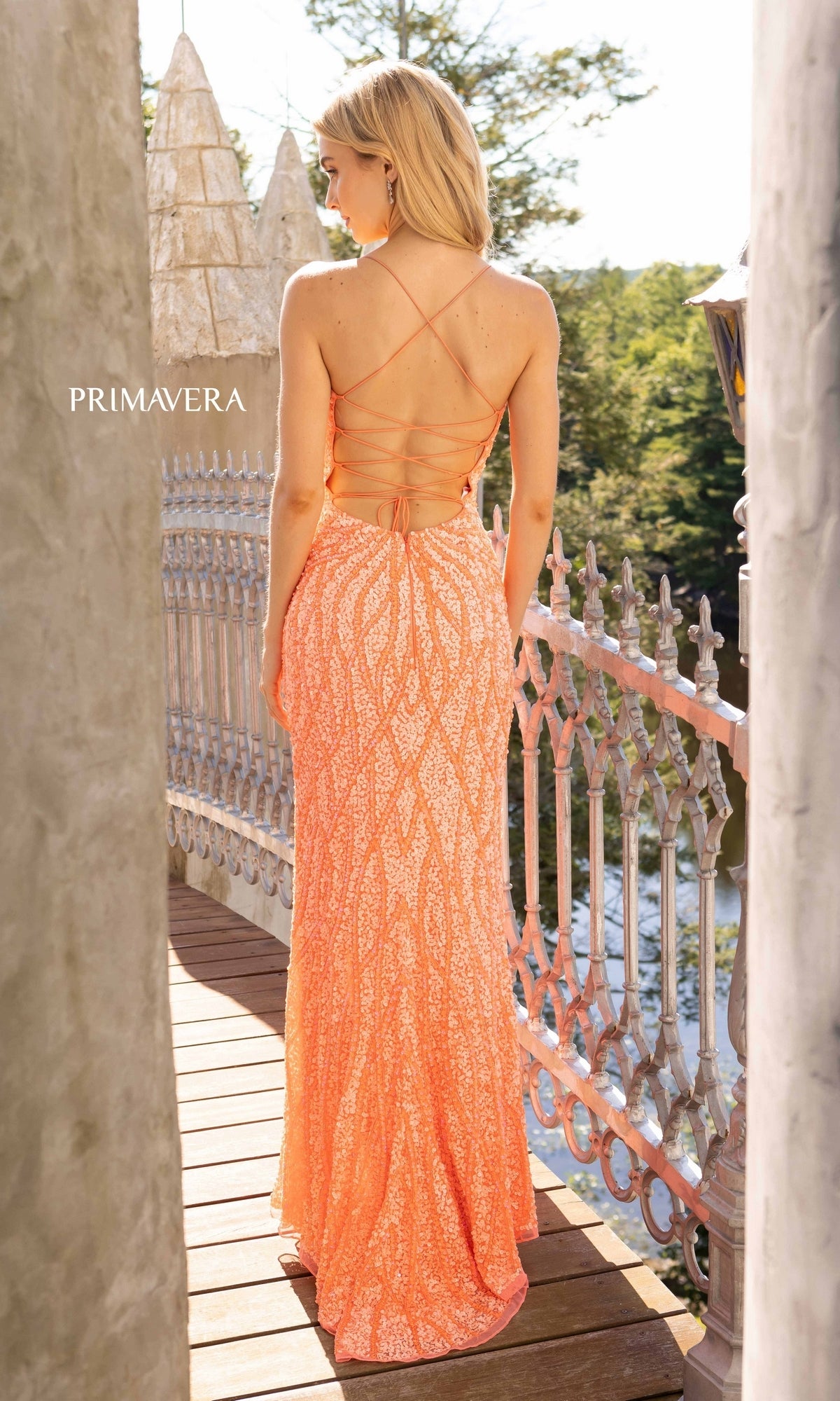 A woman in the Long Beaded Primavera Prom Gown 3959, featuring an open back, stands on a balcony overlooking water and trees.